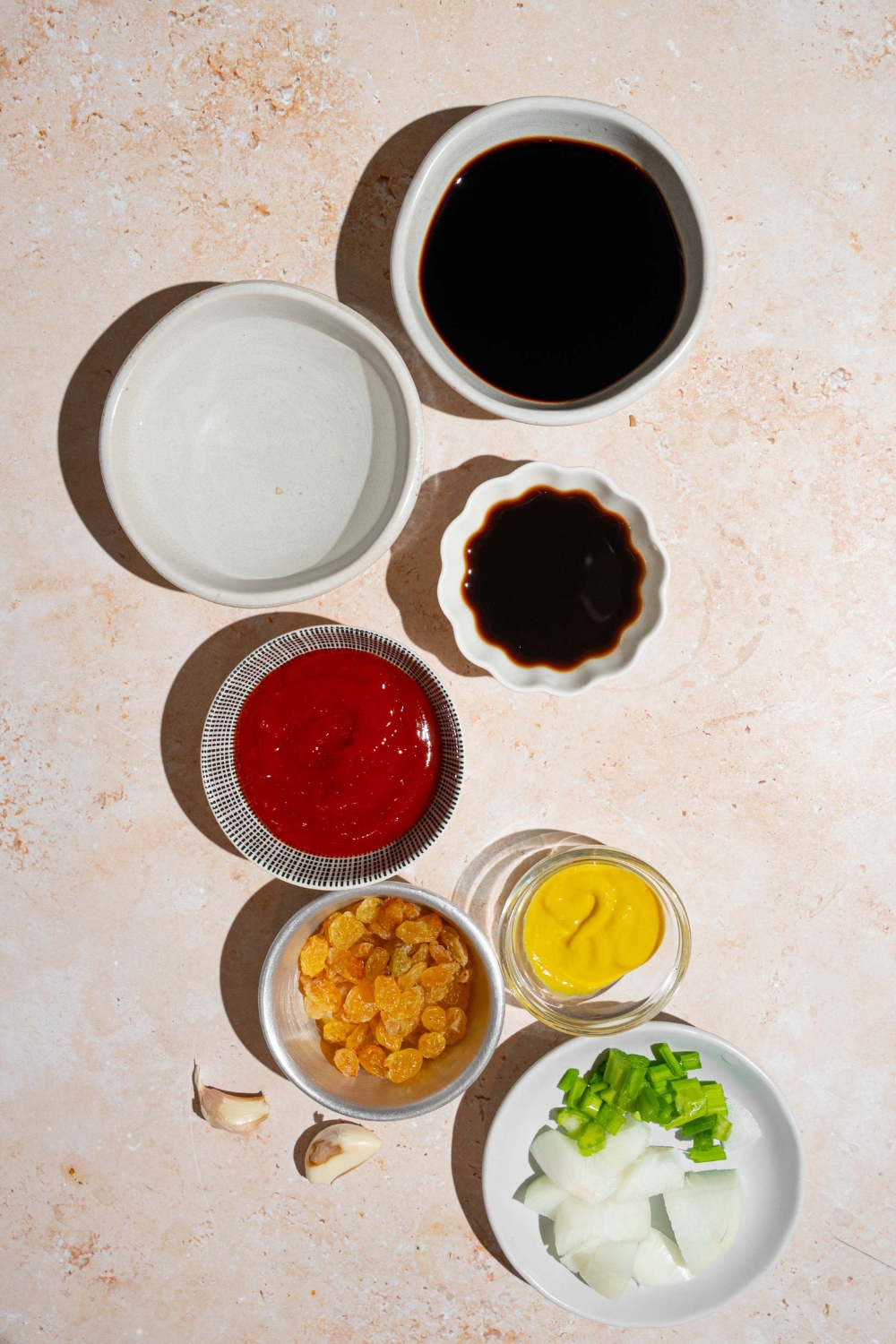 An overhead shot of several bowls in various sizes containing ingredients to make A1 steak sauce including water, balsamic vinegar, worcestershire sauce, ketchup, dijon mustard, raisins, celery, yellow onion, and seasonings.