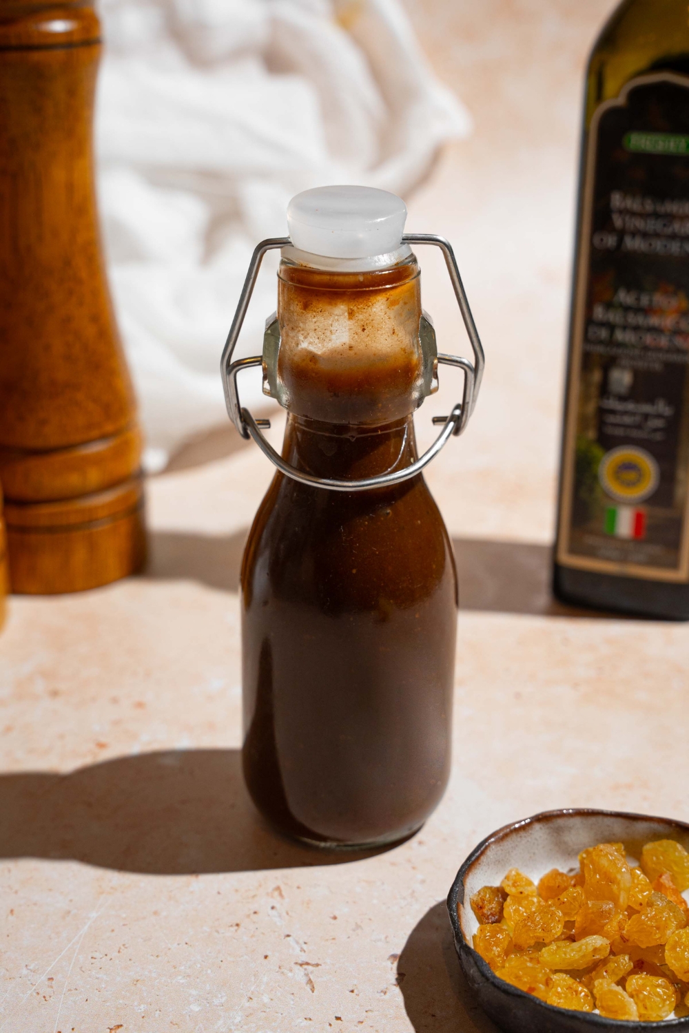 A sealed condiment jar of homemade A1 steak sauce. The jar is on a tan counter with a small plate of raisins, jar of balsamic vinegar, and a white cloth napkin.