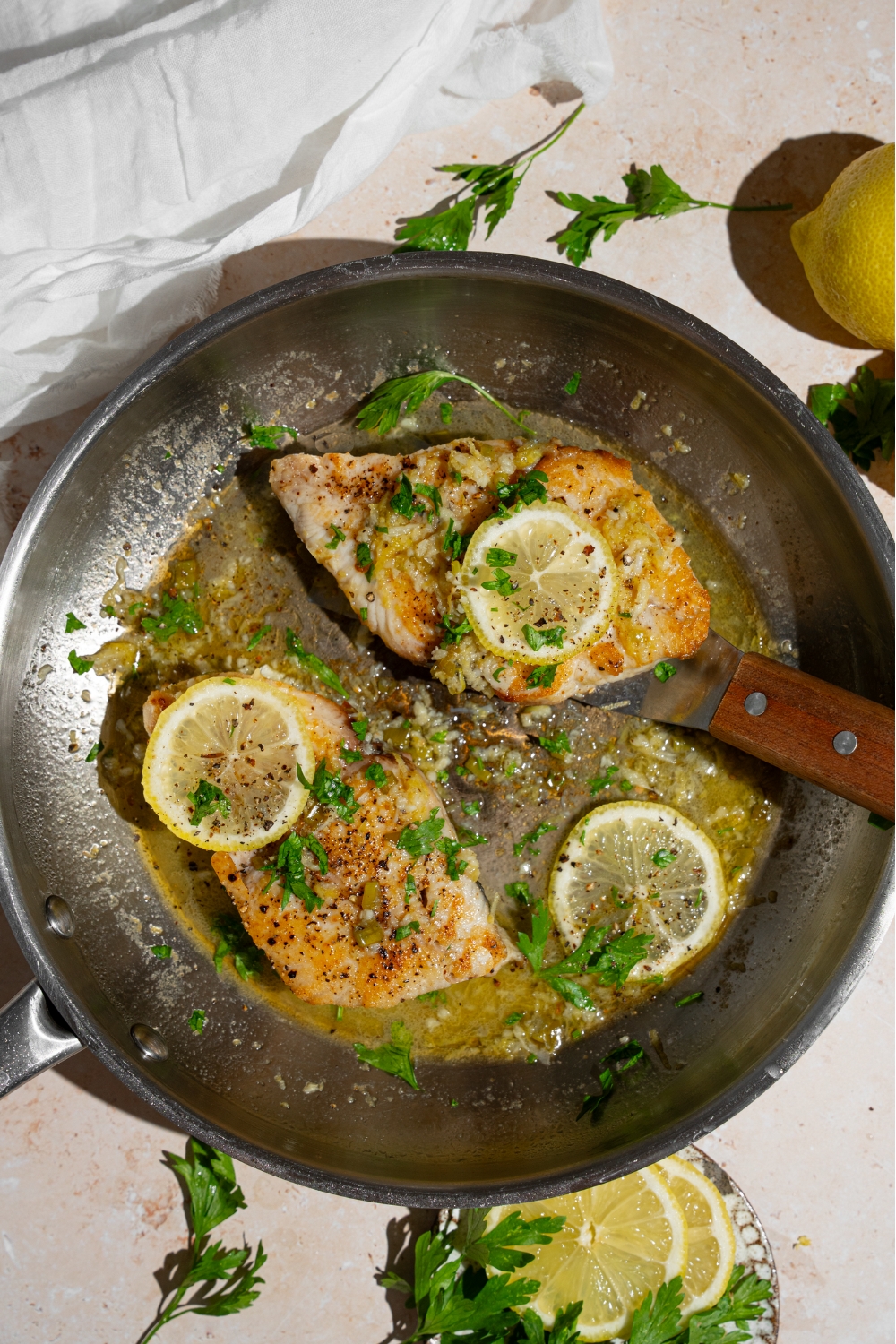Two air fryer swordfish fillets topped with lemon in a skillet with lemon garlic sauce. There is a fish spatula under one of the fillets. The skillet is on a tan counter with a plate of garnishes and white cloth napkin.