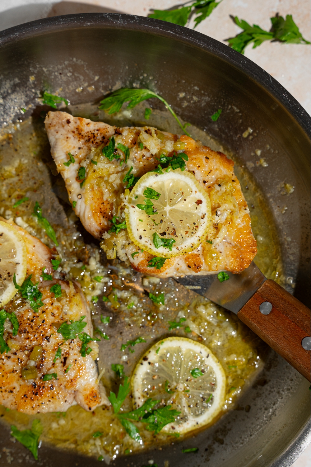 Two air fryer swordfish fillets topped with lemon in a skillet with lemon garlic sauce. There is a fish spatula under one of the fillets.