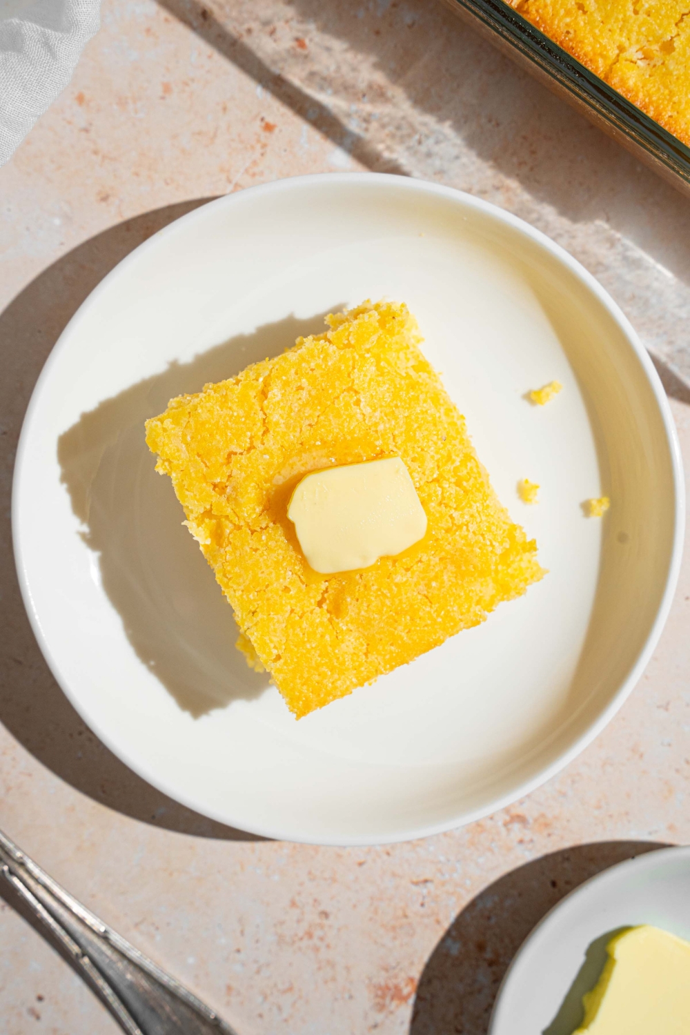 A slice of Fat Daddy's cornbread topped with a pat of butter. The corbread is on a white plate on a tan counter with a small plate of butter.