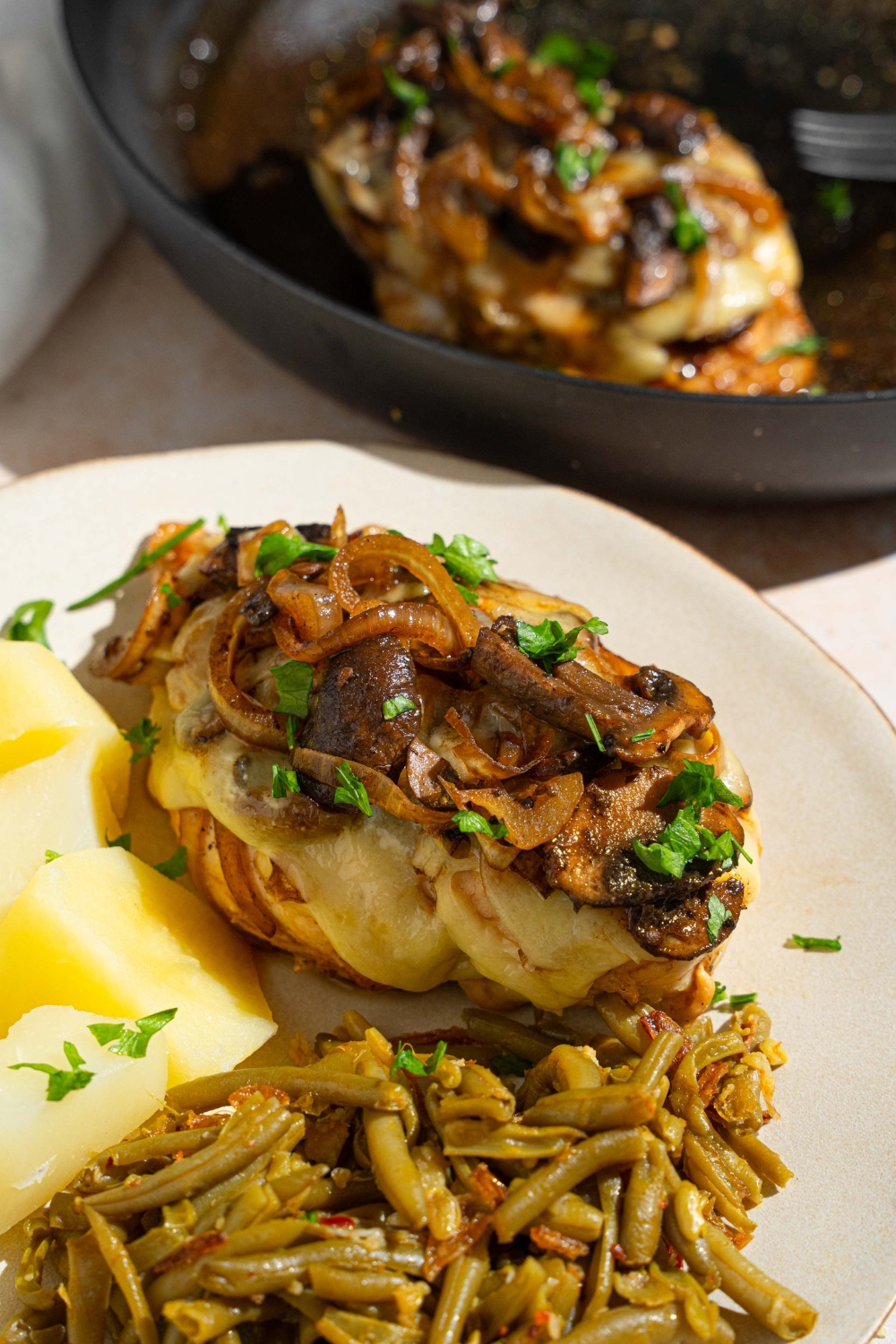 A piece of Texas Roadhouse smothered chicken on a white plate with cubed potatoes and green beans. The plate is on garnished with fresh parsley. The plate is on a tan counter with a skillet with additional chicken.