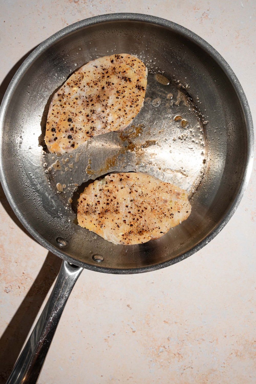 A skillet with two swordfish fillets searing in oil. The skillet is on a tan counter.