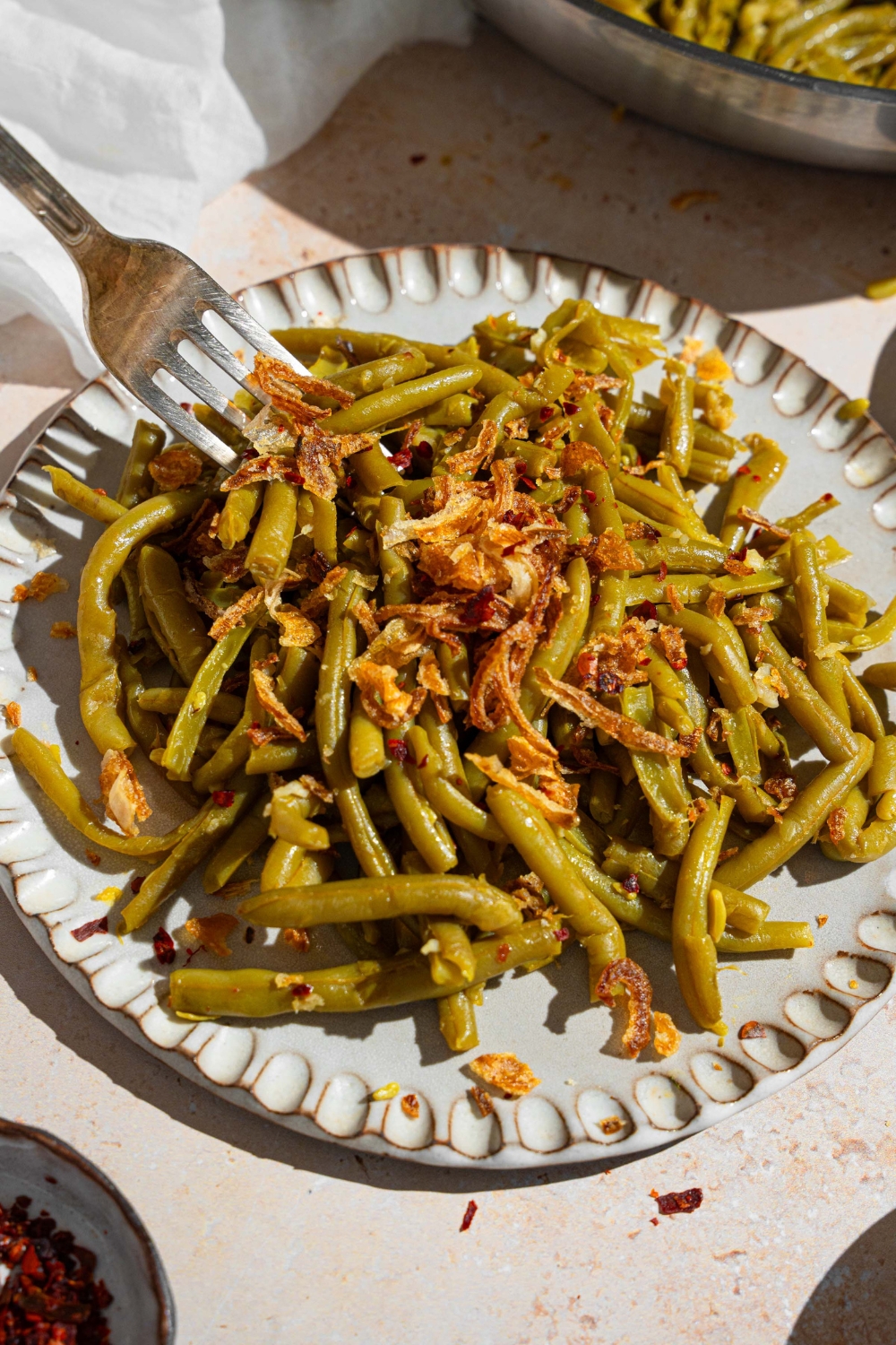 Canned green beans cooked in butter and spices and topped with crispy onion strips. A fork is taking a bite. The green beans are on a ceramic plate on a tan counter with a white cloth napkin and small ramekin of red pepper flakes.
