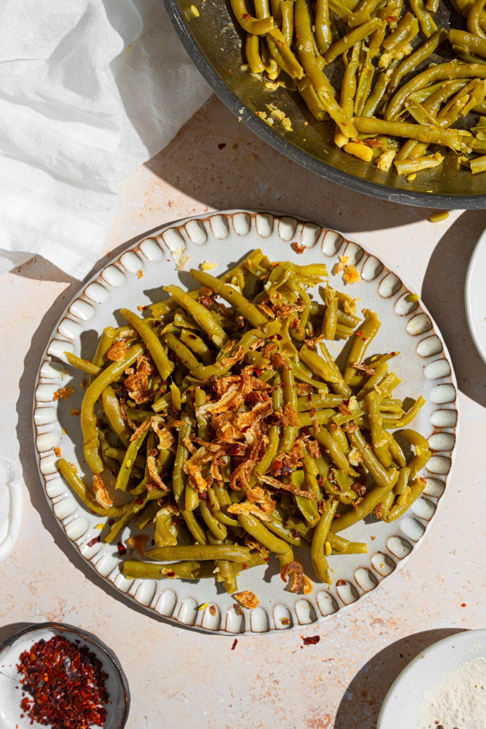 Canned green beans cooked in butter and spices and topped with crispy onion strips. The green beans are on a ceramic plate on a tan counter with a skillet of green beans and small ramekin of red pepper flakes.
