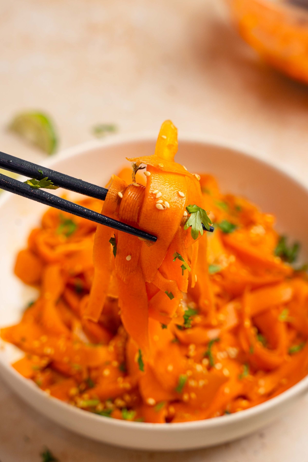 A close up of a pair of chopsticks holding a bite of carrot salad garnished with sesame seeds and cilantro. There is a bowl of carrot salad blurred in the background.