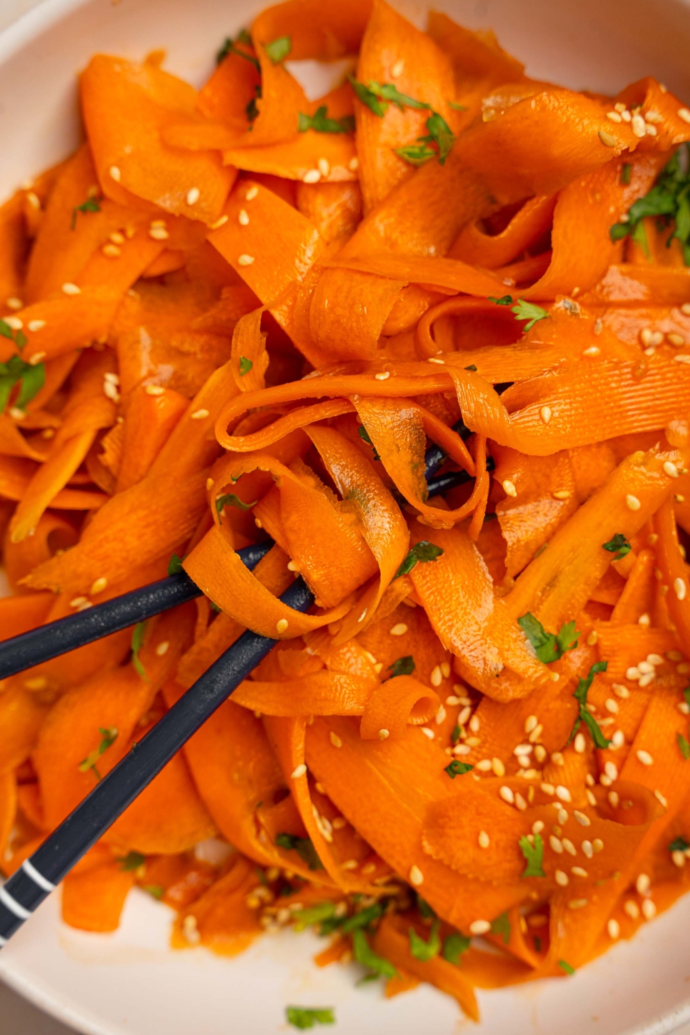 A bowl of carrot salad garnished with sesame seeds and fresh cilantro. There is a pair of chopsticks in the bowl.