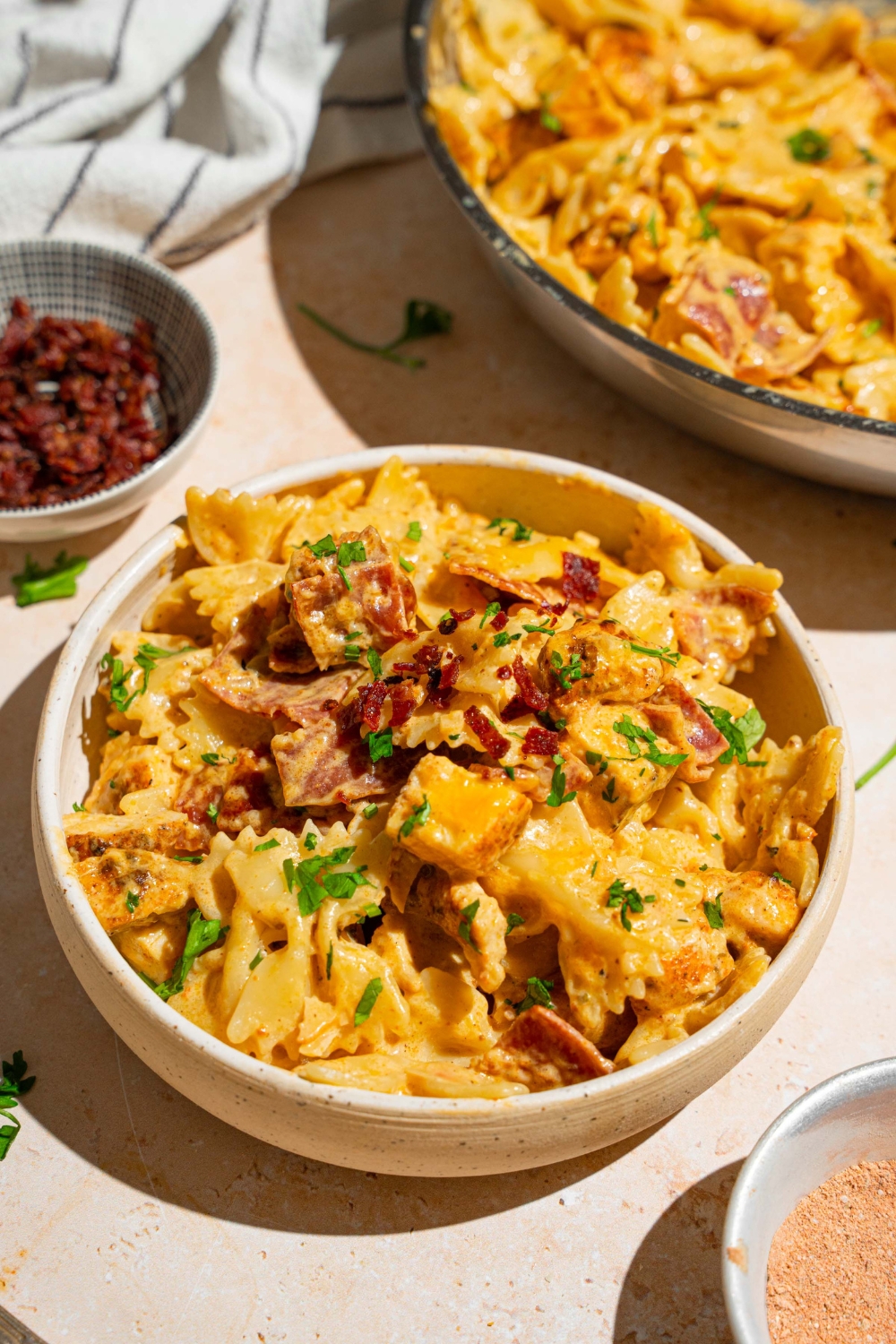 A white bowl with chicken bacon ranch pasta garnished with fresh parsley. The bowl is on a tan counter with a skillet of pasta and small bowl of bacon.