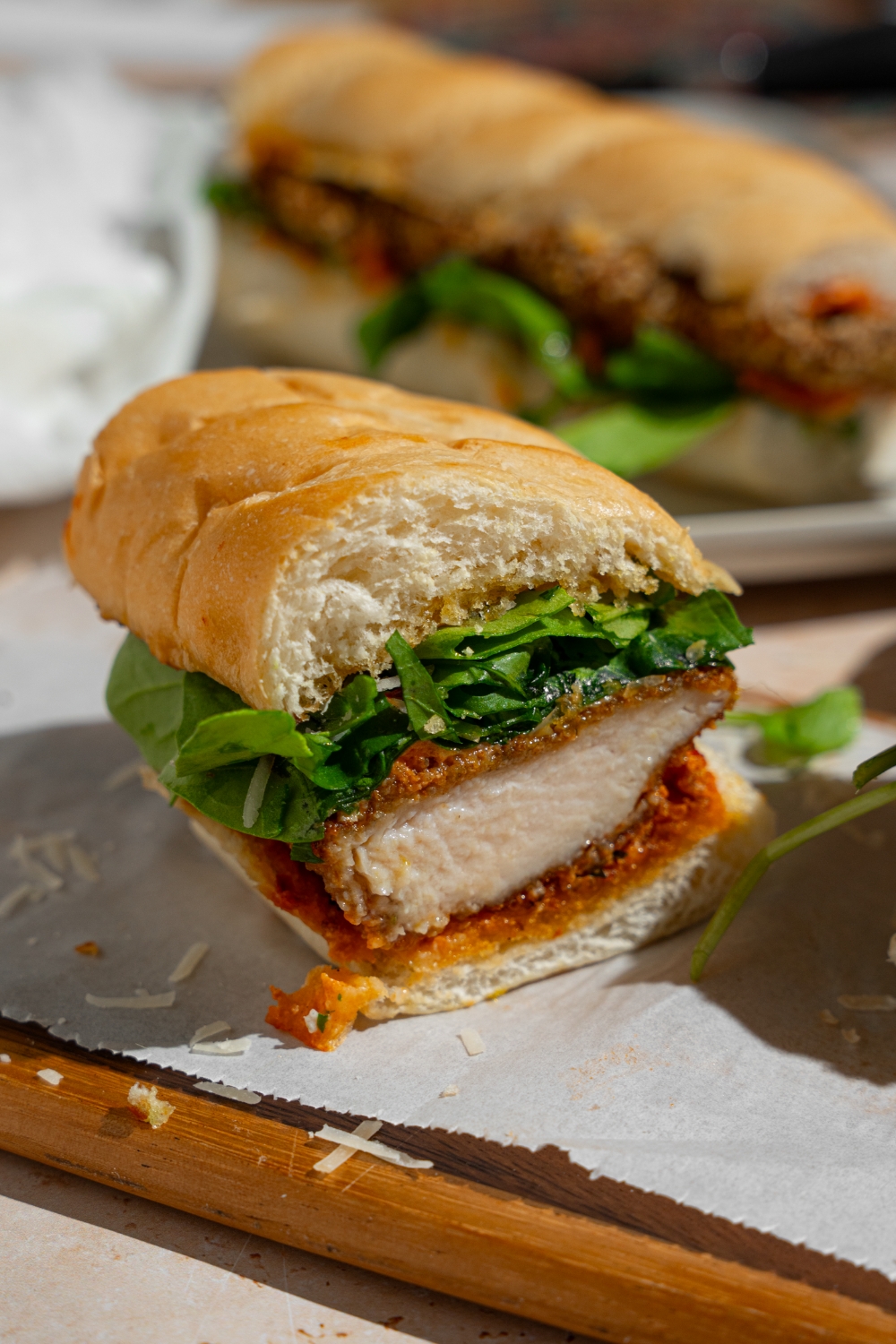Half of a chicken cutlet sandwich on a wooden board lined with parchment paper. The sandwich shows a breaded chicken cutlet, arugula, and creamy tomato sauce in between sliced garlic bread. The board is on a tan counter with an additional whole sandwich.