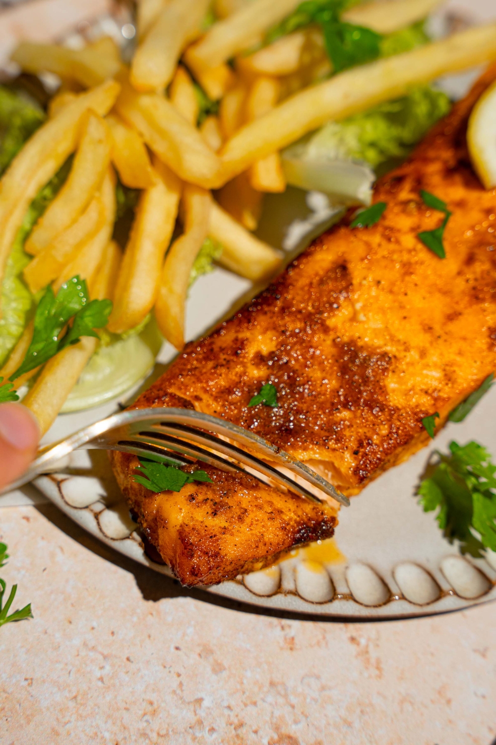 A ceramic plate with frozen air fryer salmon served with lettuce and french fries and garnished with fresh parsley and lemon. A fork is taking a bite of salmon.
