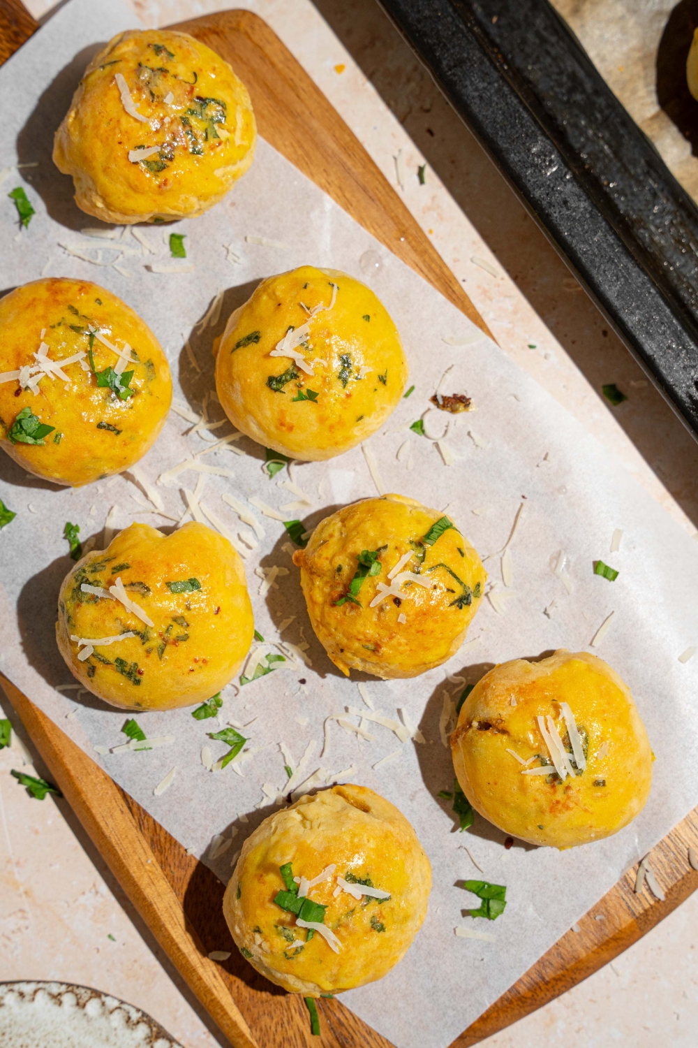 A wooden board lined with parchment paper with several garlic parmesan cheeseburger bombs garnished with fresh parsley and grated cheese. The board is on a tan counter.