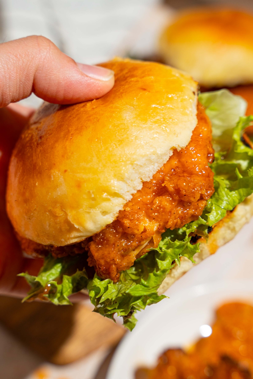 A close up of a hand holding a hot honey chicken sandwich with lettuce. There is a board of sandwiches blurred in the background.