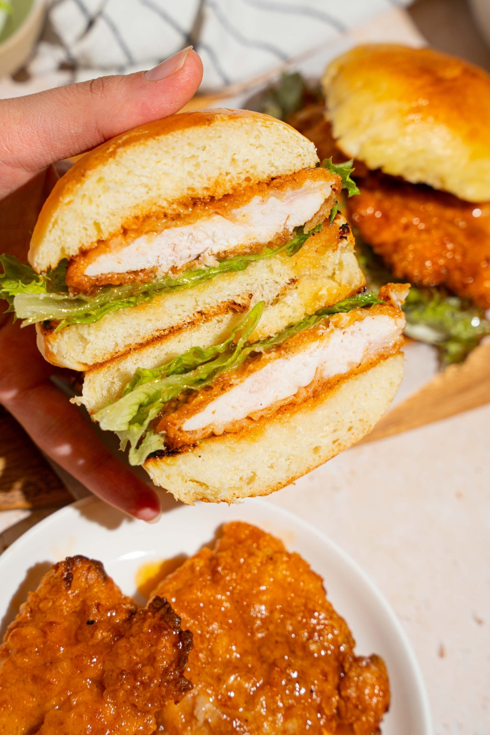 A hand holding a hot honey chicken sandwich sliced in half. There is a board of sandwiches and plate of hot honey chicken in the background.