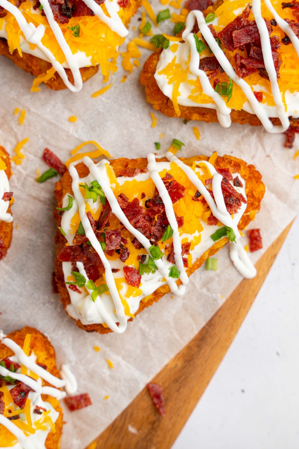 Several loaded hashbrowns topped with sour cream mixture, cheddar cheese, and crumbled bacon on a wooden board lined with parchment paper. The hashbrowns are drizzled with sour cream and garnished with sliced chives. The board is on a white counter.