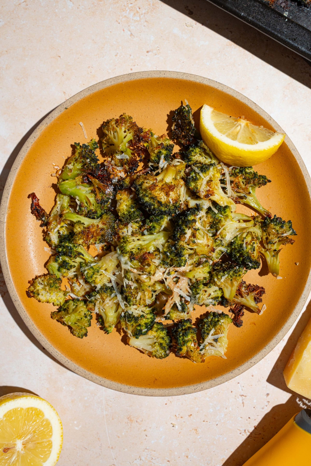 An orange plate with roasted frozen broccoli garnished with grated parmesan and lemon. The plate is on a tan counter with a lemon.