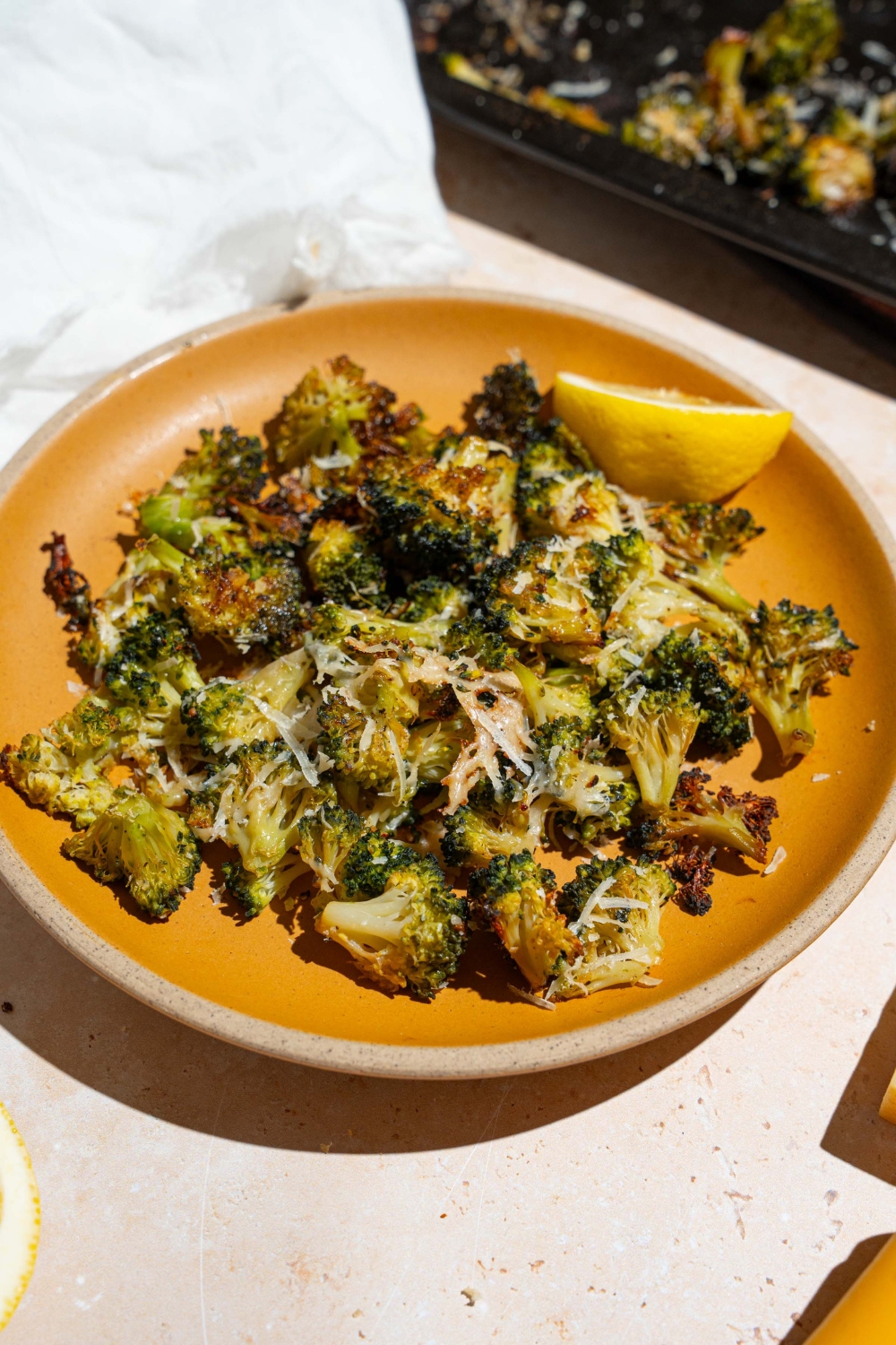 An orange plate with roasted frozen broccoli garnished with grated parmesan and lemon. The plate is on a tan counter with a white cloth napkin.