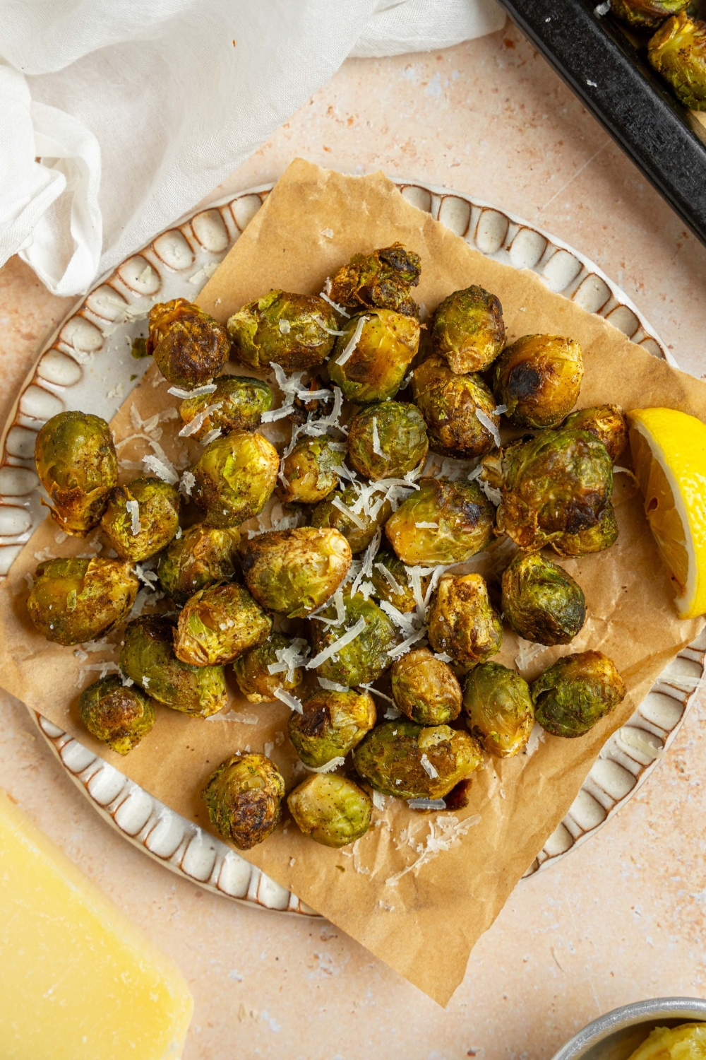 A ceramic plate lined with parchment paper with roasted frozen brussel sprouts garnished with grated parmesan cheese and lemon. The plate is on a tan counter with a baking sheet of brussel sprouts and white cloth napkin.
