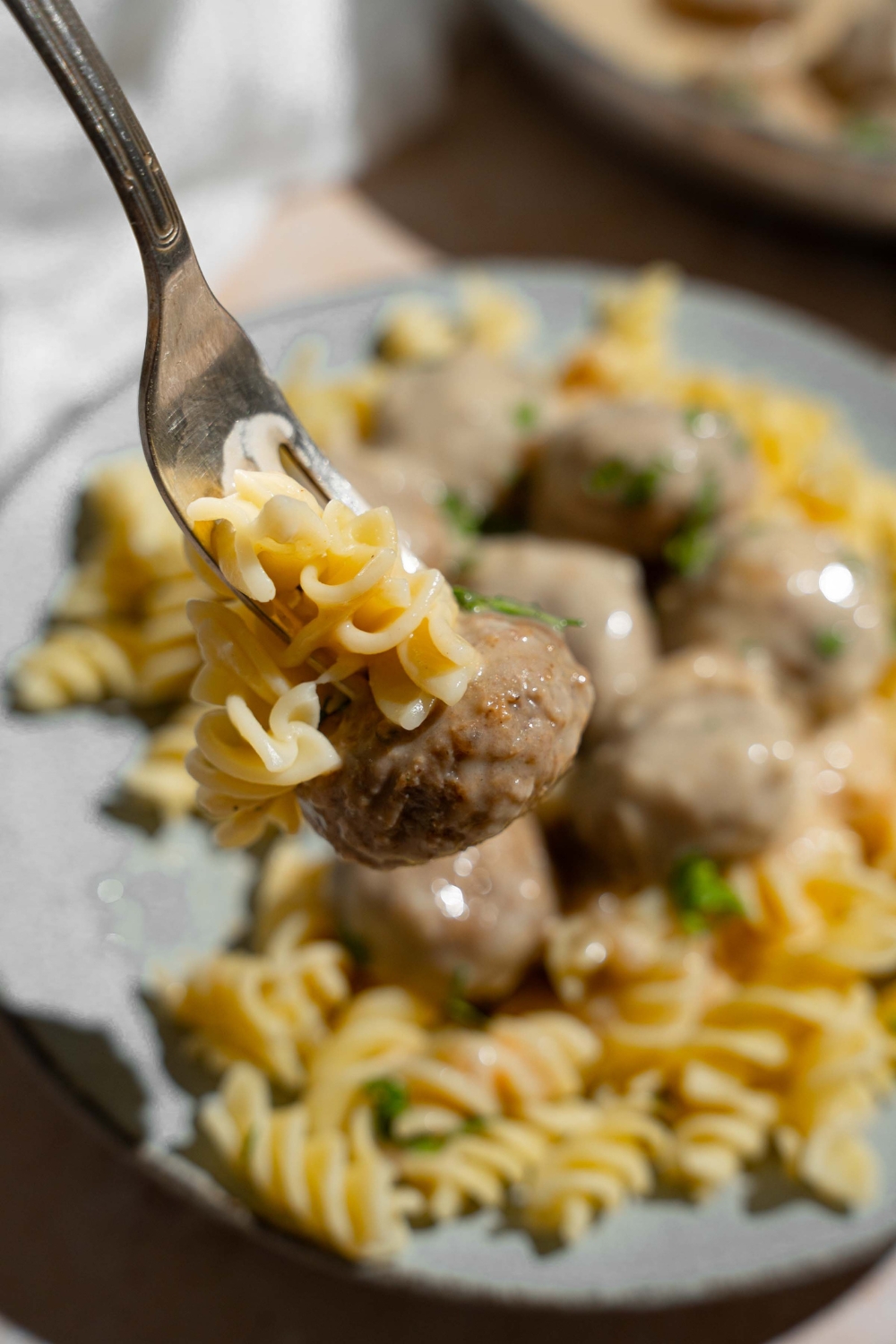 A close up of a fork with a bite of pasta and swedish meatball. There is a plate of pasta and meatballs blurred in the background.