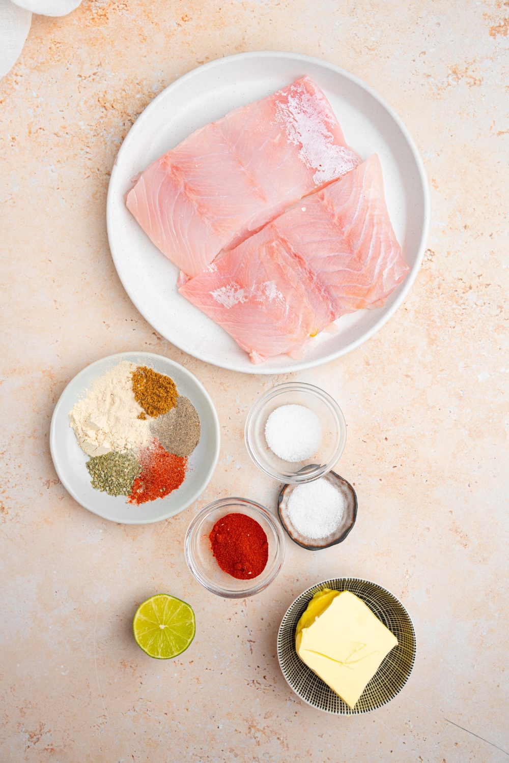 An overhead shot of several bowls in various sizes containing various ingredients to make blackened grouper including grouper filets, butter, garlic, lime, sugar, and seasonings.