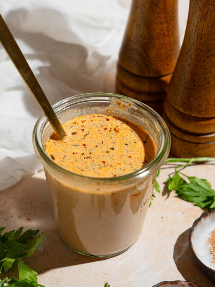 A glass jar of cajun cream sauce with a spoon in the jar. The jar is on a tan counter with fresh parsley, salt and pepper mills, and white cloth napkin.