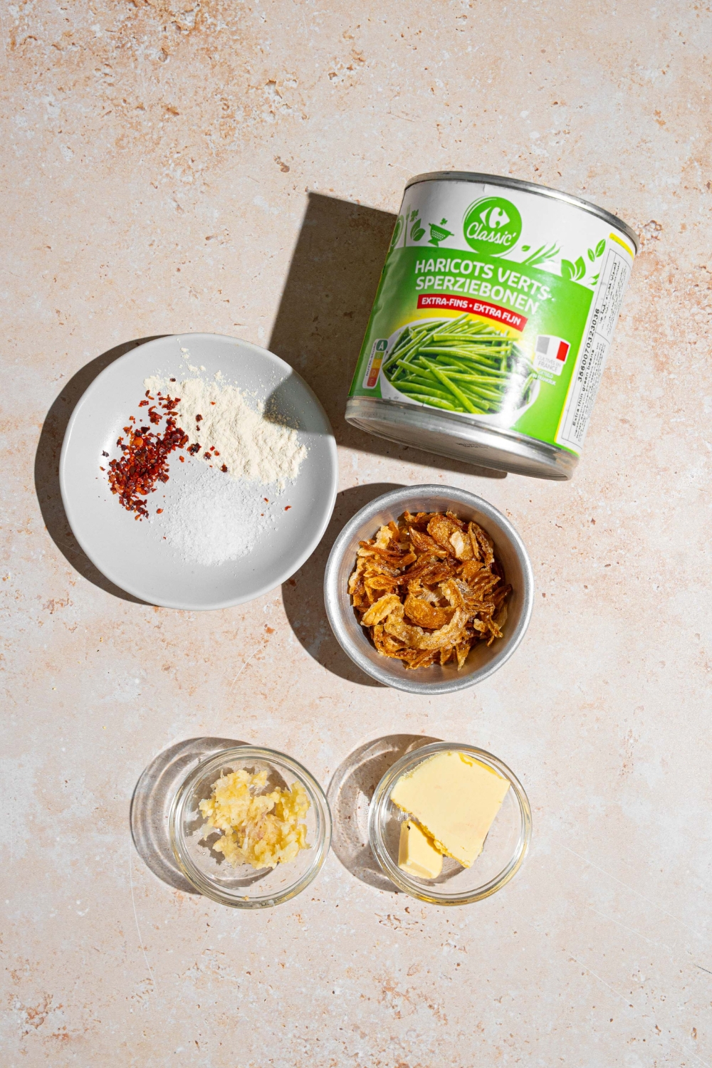 An overhead shot of several bowls in various sizes containing ingredients to make canned green beans including a can of green beans, butter, garlic, red pepper flakes, and seasonings.