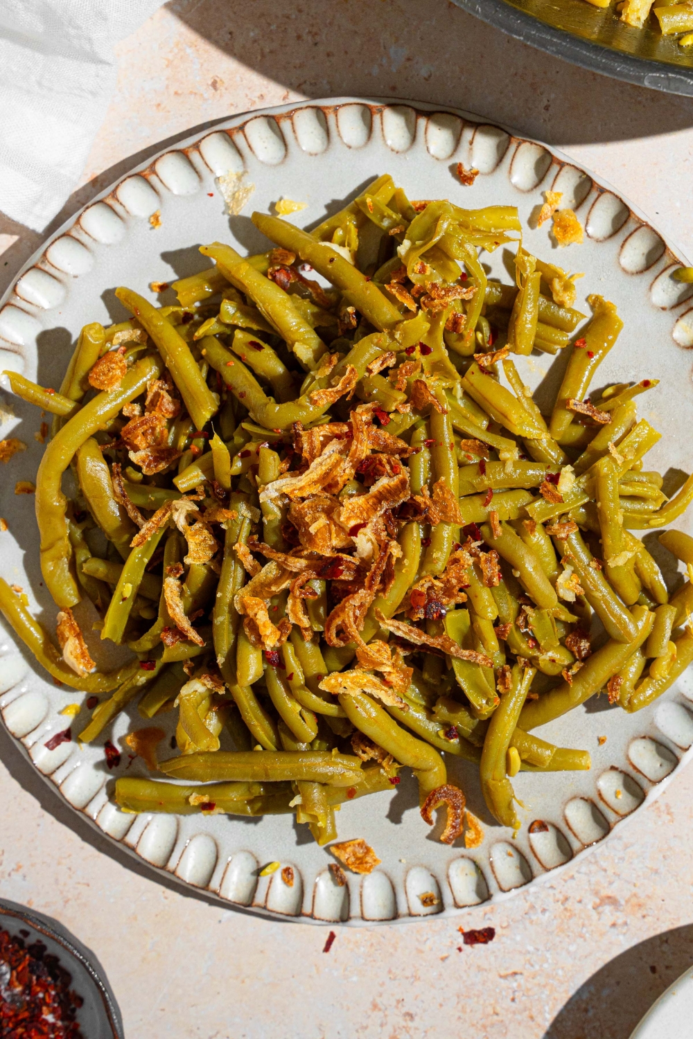 Canned green beans cooked in butter and spices and topped with crispy onion strips. The green beans are on a ceramic plate on a tan counter with a skillet of green beans and small ramekin of red pepper flakes.