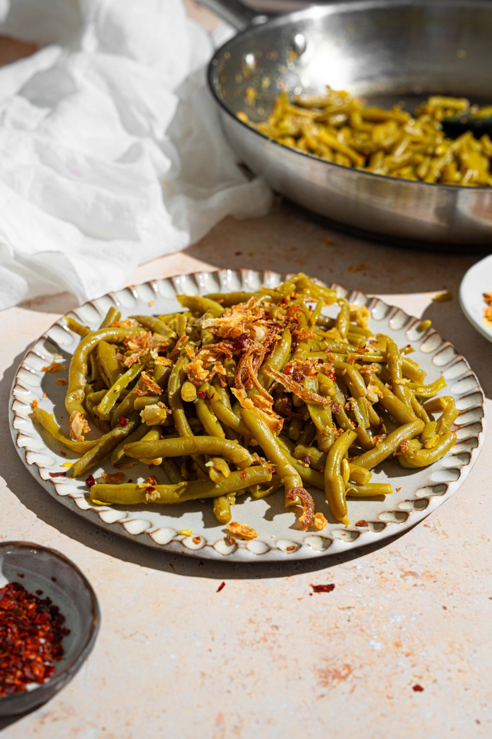 Canned green beans cooked in butter and spices and topped with crispy onion strips. The green beans are on a ceramic plate on a tan counter with a skillet of green beans and small ramekin of red pepper flakes.