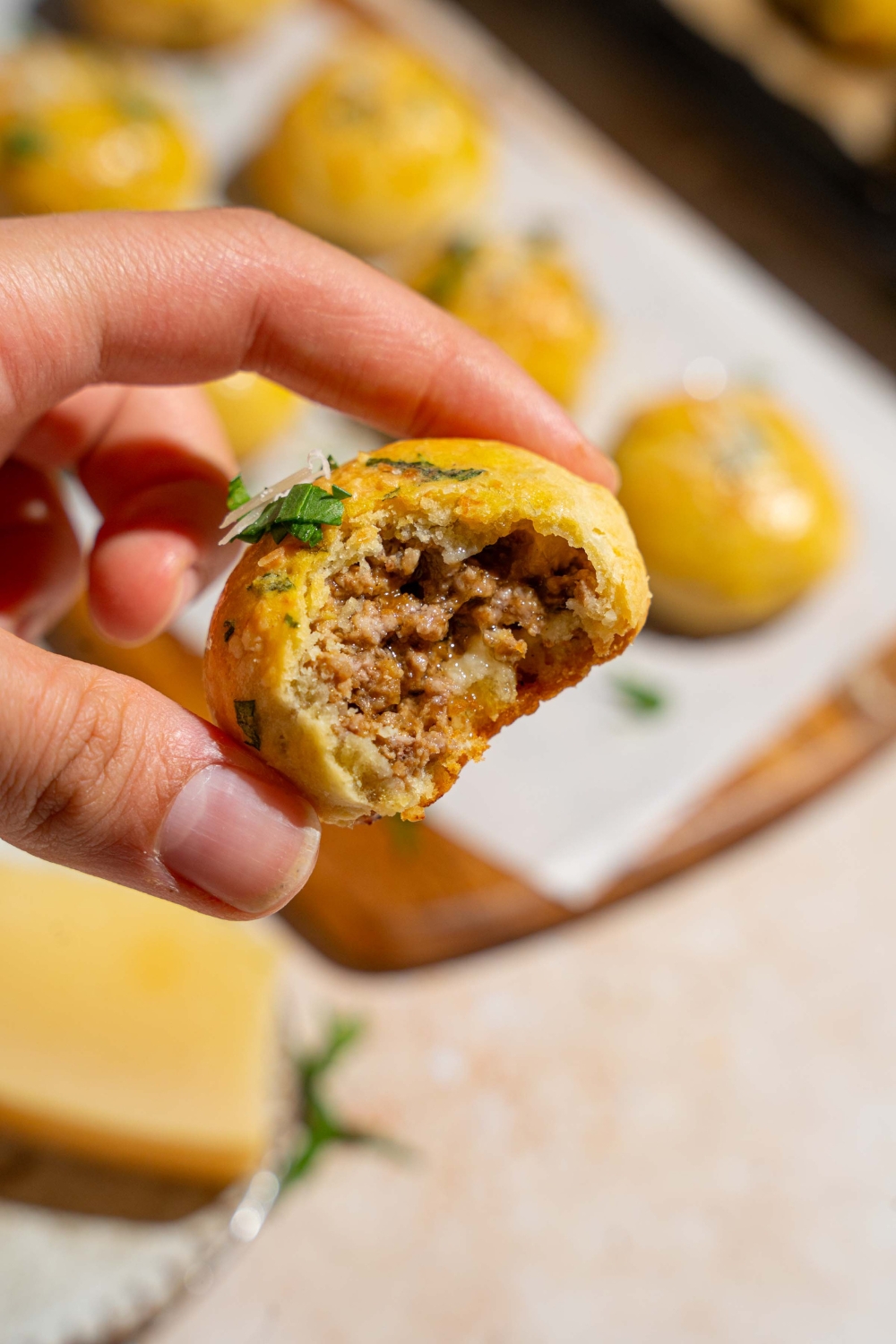 A close up of a hand holding a bitten garlic parmesan cheeseburger bite garnished with fresh parsley and grated cheese. There is a board of cheeseburger bites blurred in the background.