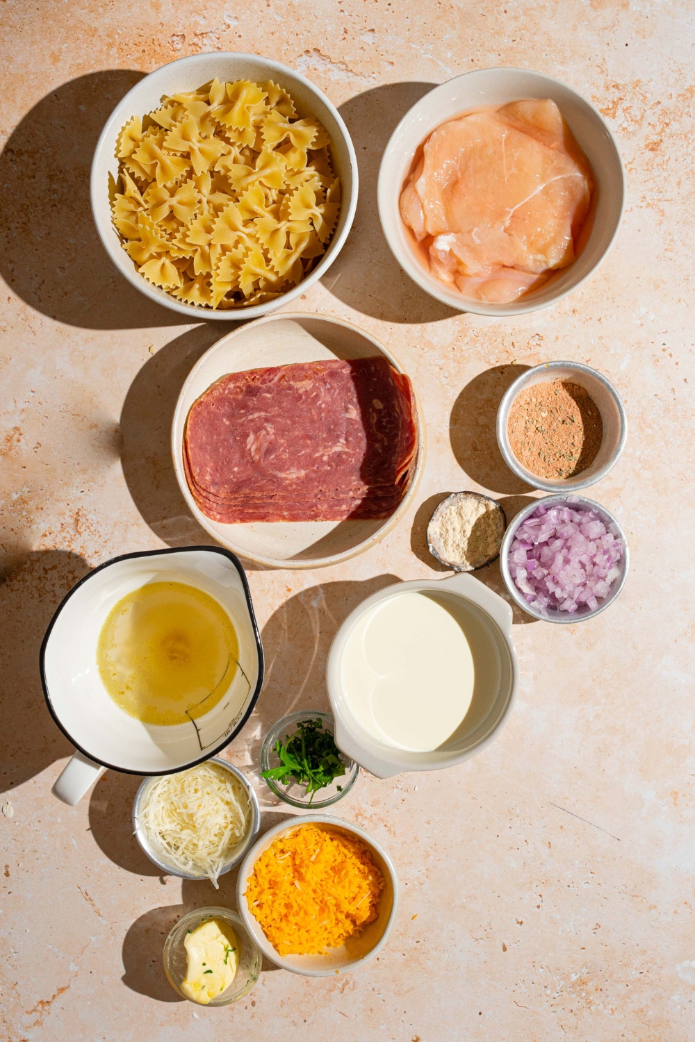 An overhead shot of several bowls in various sizes containing ingredients to make chicken bacon ranch pasta including chicken, pasta, bacon, cheddar cheese, shallots, chicken broth, cream, butter, and seasonings.