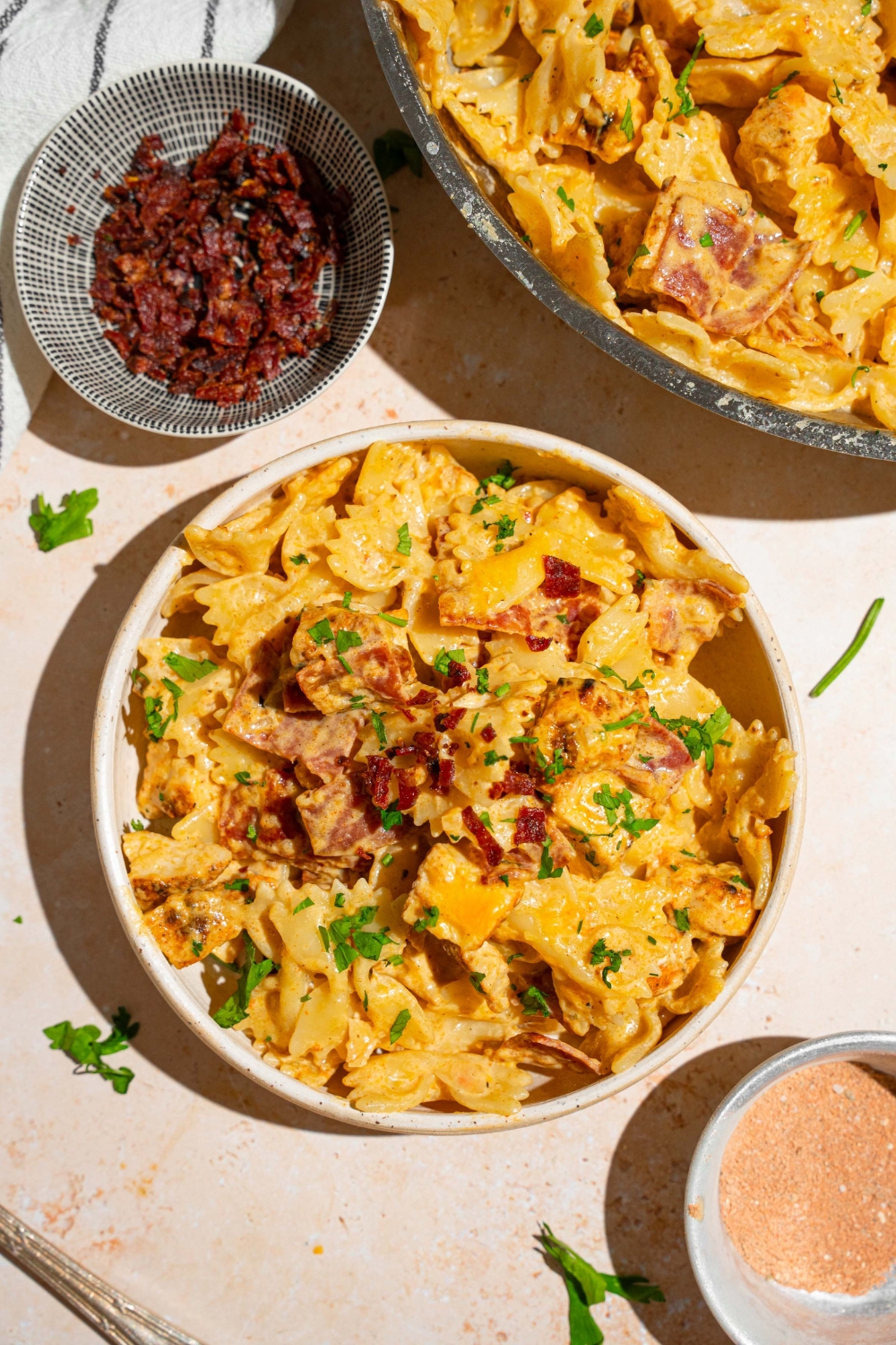 A white bowl with chicken bacon ranch pasta garnished with fresh parsley. The bowl is on a tan counter with a skillet of pasta and small bowl of bacon.