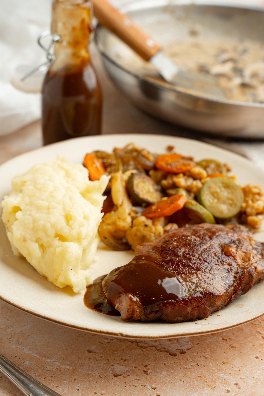 A white plate with mashed potatoes, roasted vegetables, and steak topped with homemade A1 steak sauce. The plate is on a tan counter with a condiment jar of steak sauce and skillet.