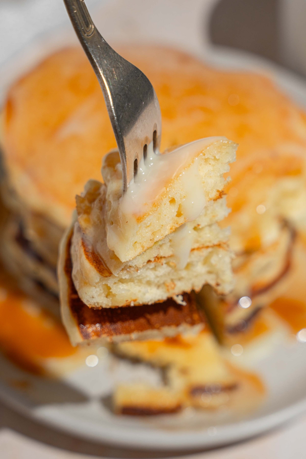 A close up of a fork with a bite of tres leches pancakes. There is a plate of pancakes blurred in the background.