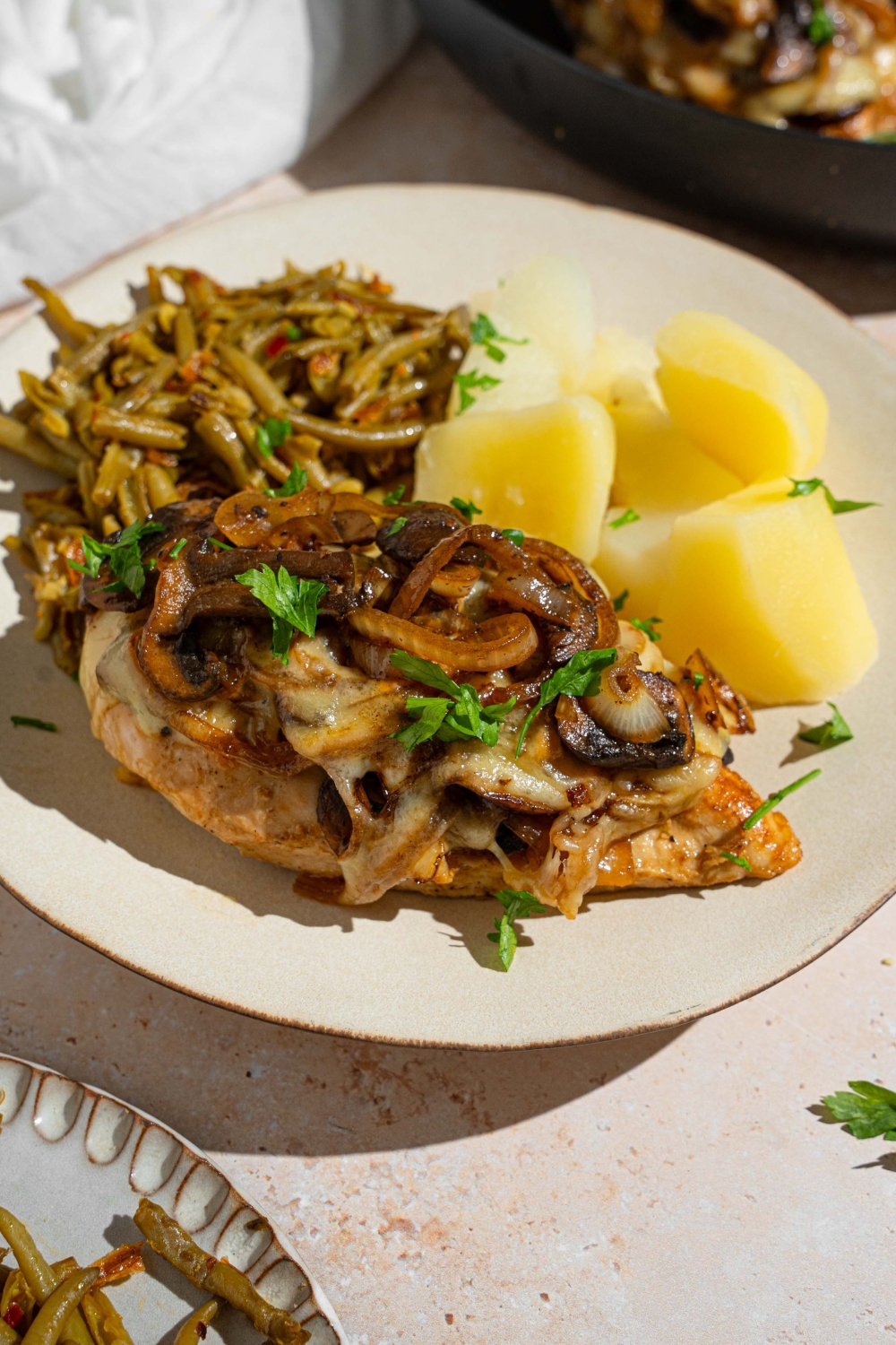 A piece of Texas Roadhouse smothered chicken on a white plate with cubed potatoes and green beans. The plate is on garnished with fresh parsley. The plate is on a tan counter with a skillet with additional chicken.