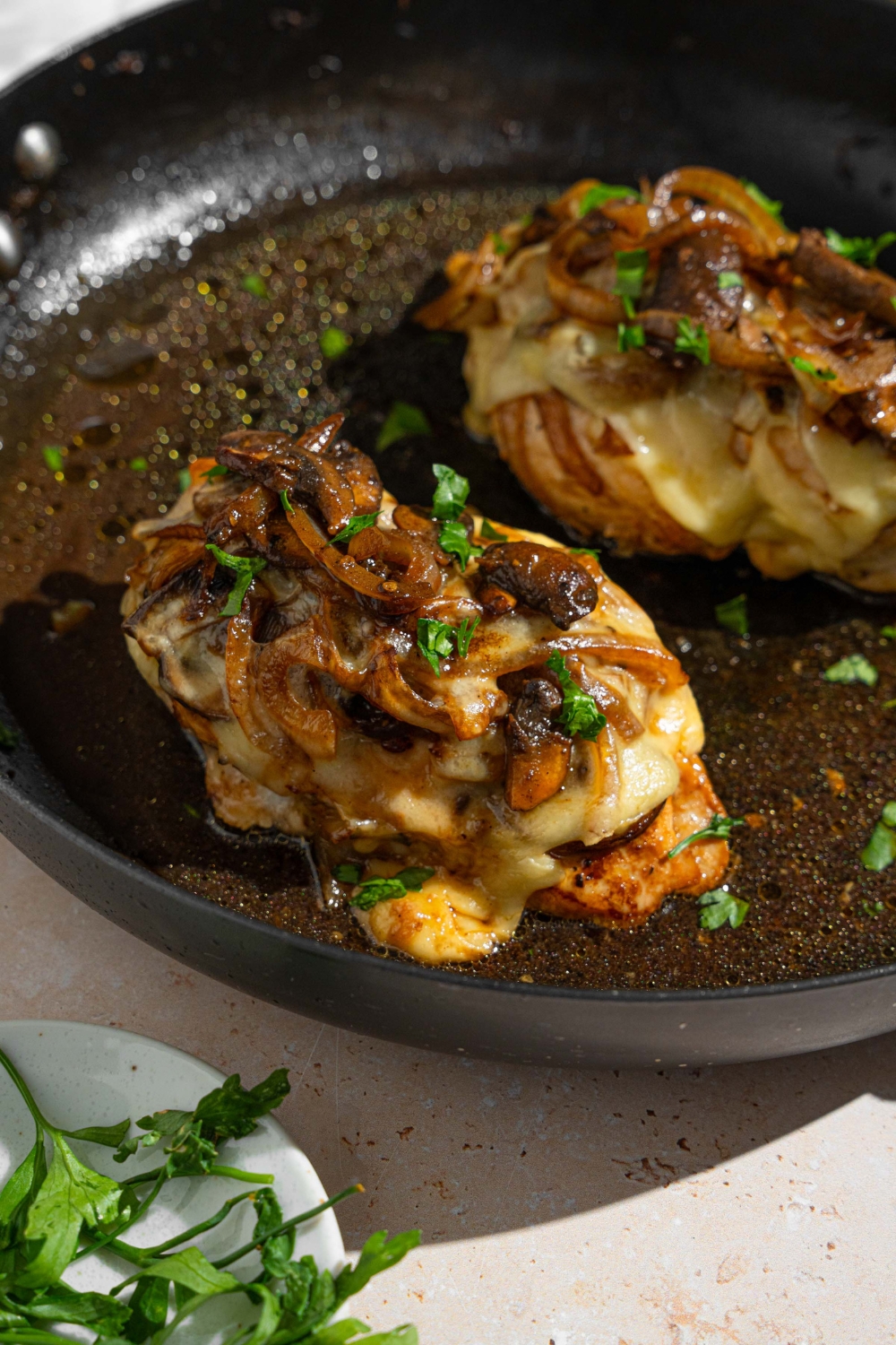 Two Texas Roadhouse smothered chicken breasts on a skillet in juices and garnished with fresh parsley. The skillet is on a tan counter with a small plate of garnishes.