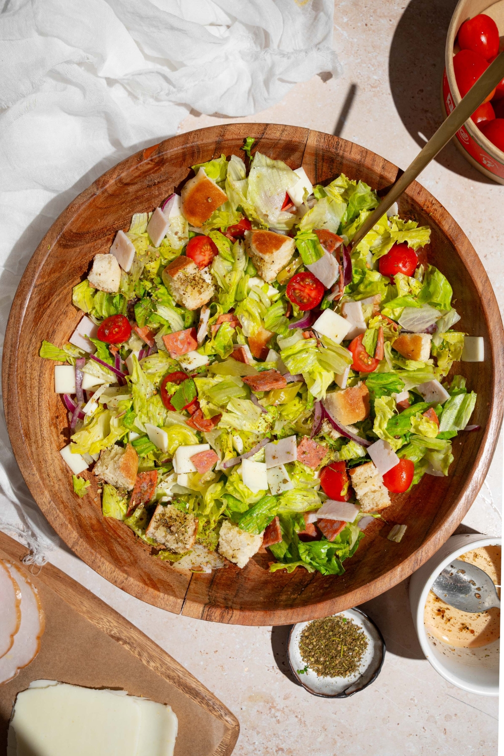 A wooden salad bowl of copycat Jersey Mike's sub in a tub including mixed lettuce, tomatoes, provolone cheese, diced meats, and bread croutons. There is a fork in the salad. The bowl is on a tan counter with a small ramekin of seasonings and dressing.