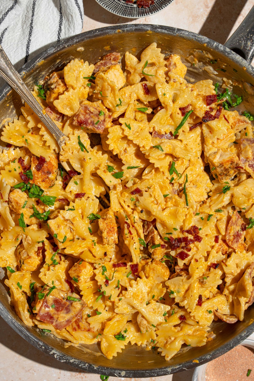 A skillet with chicken bacon ranch pasta garnished with fresh parsley. There is a fork in the skillet. The skillet is on tan counter with a striped napkin.