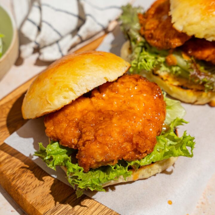 A wooden board lined with parchment paper with two hot honey chicken sandwiches. The board is on a tan counter with a white striped napkin.