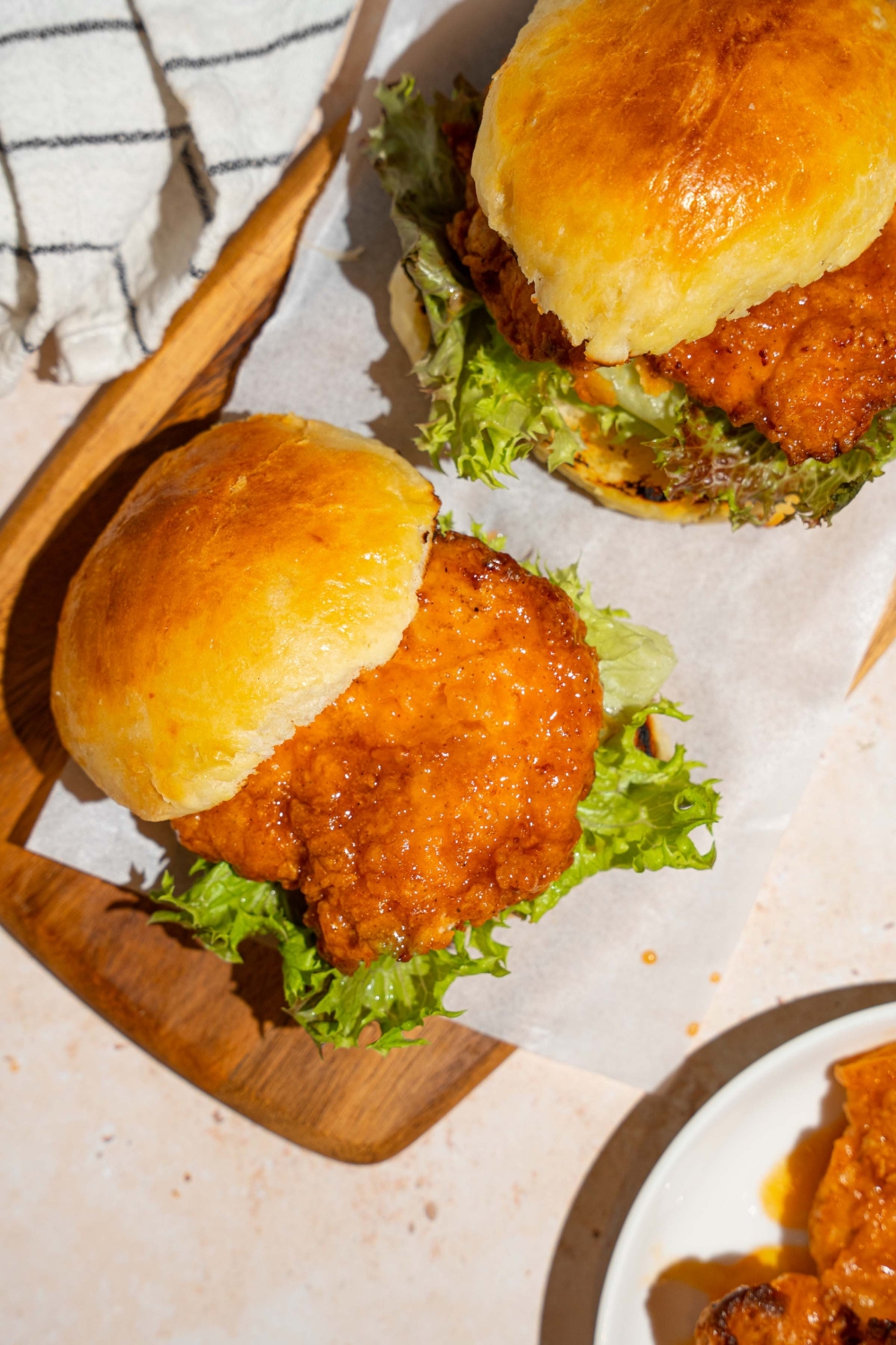 A wooden board lined with parchment paper with two hot honey chicken sandwiches. The board is on a tan counter with a white striped napkin.