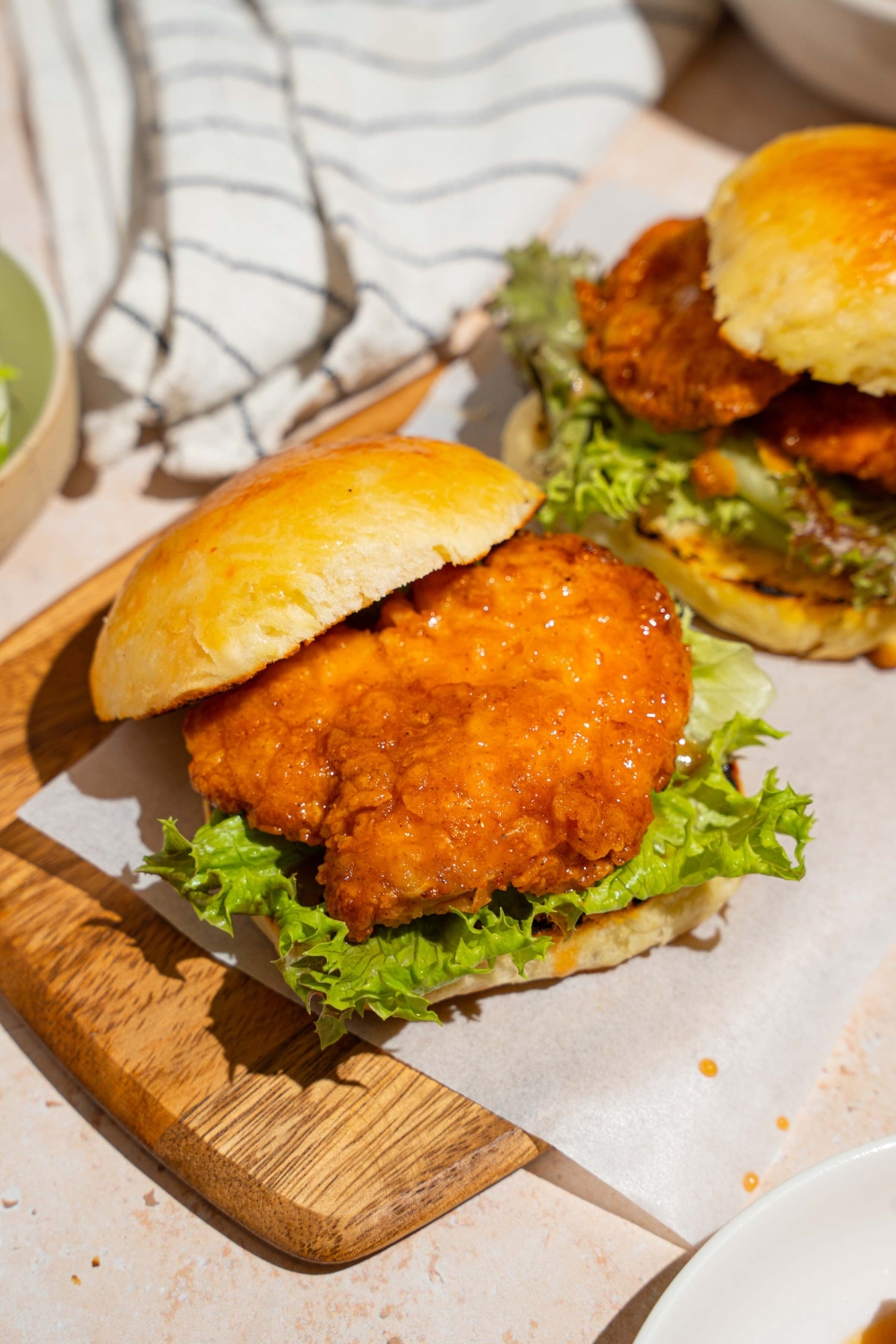 A wooden board lined with parchment paper with two hot honey chicken sandwiches. The board is on a tan counter with a white striped napkin.
