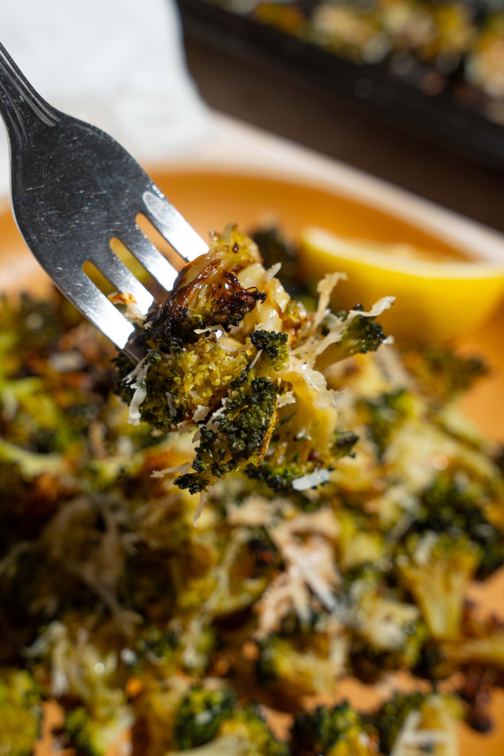 A close up of a fork with a bite of roasted frozen broccoli. There is a plate of broccoli blurred in the background.