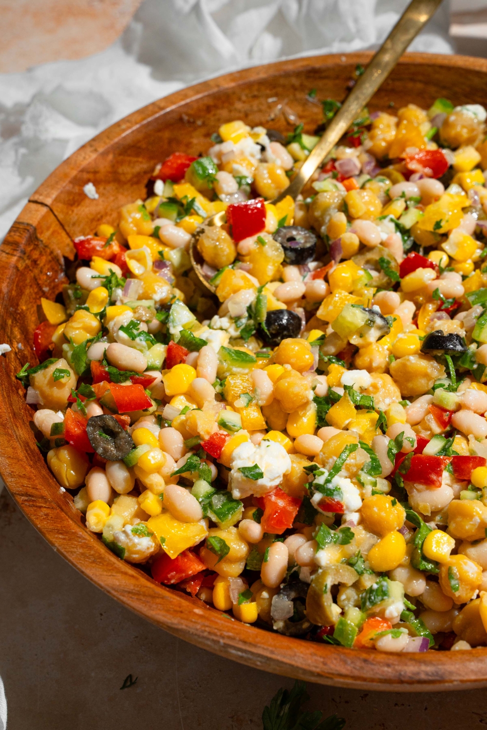 A wooden bowl with Mediterranean dense bean salad including black olives, corn, navy beans, chickpeas, bell peppers, feta cheese, and cucumber garnished with fresh parsley. There is a spoon in the bowl. The bowl is on a tan counter with a white cloth napkin.