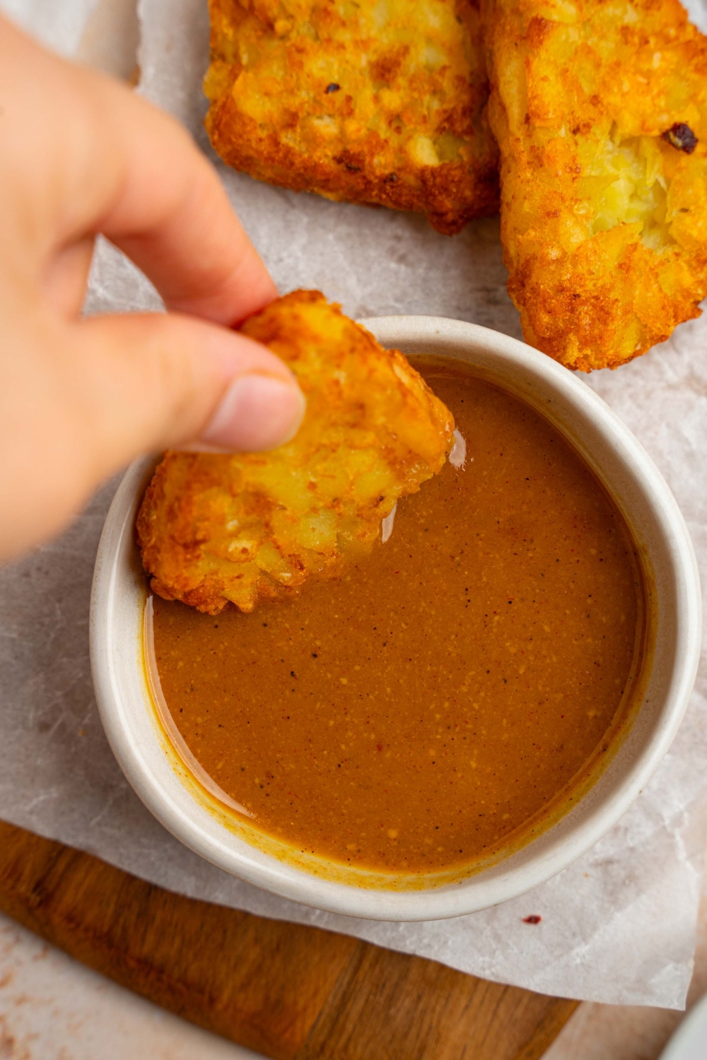 A hand dipping a crispy hashbrown into a small bowl of McDonald's golden sauce. The bowl is on a wooden board lined with parchment paper with crispy hashbrowns.
