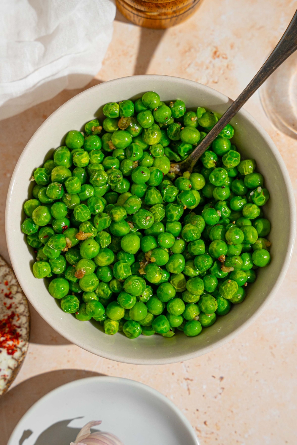 A white bowl with cooked frozen peas tossed in seasonings. There is a spoon in the bowl. The bowl is on a tan counter with a white cloth napkin.