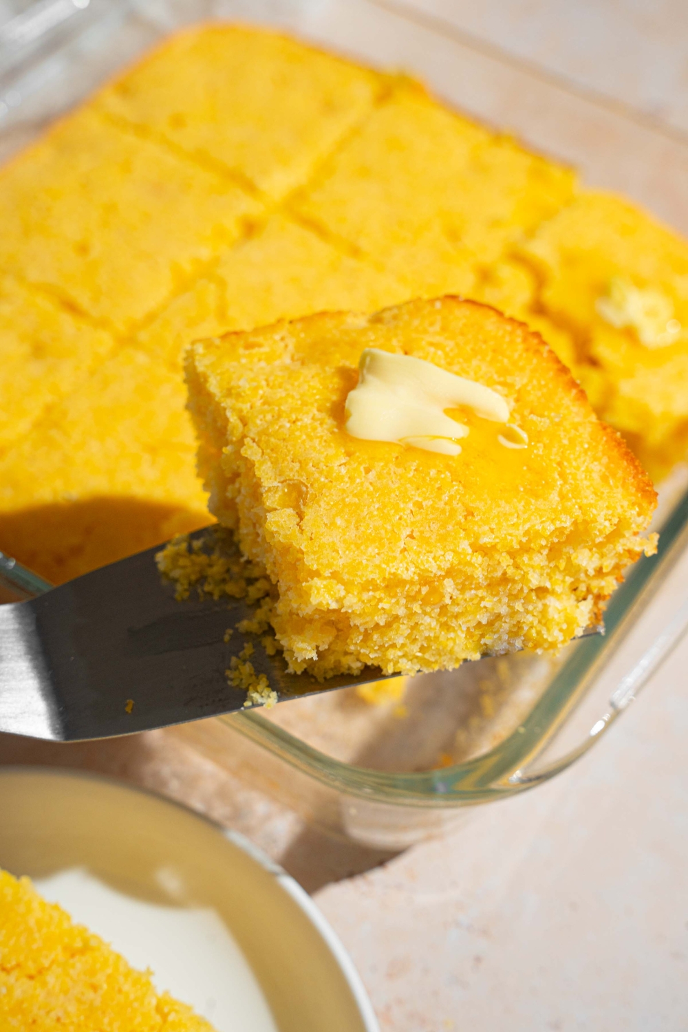 A close up of a spatula with a slice of cornbread topped with a pat of butter. There is a glass dish of cornbread blurred in the background.