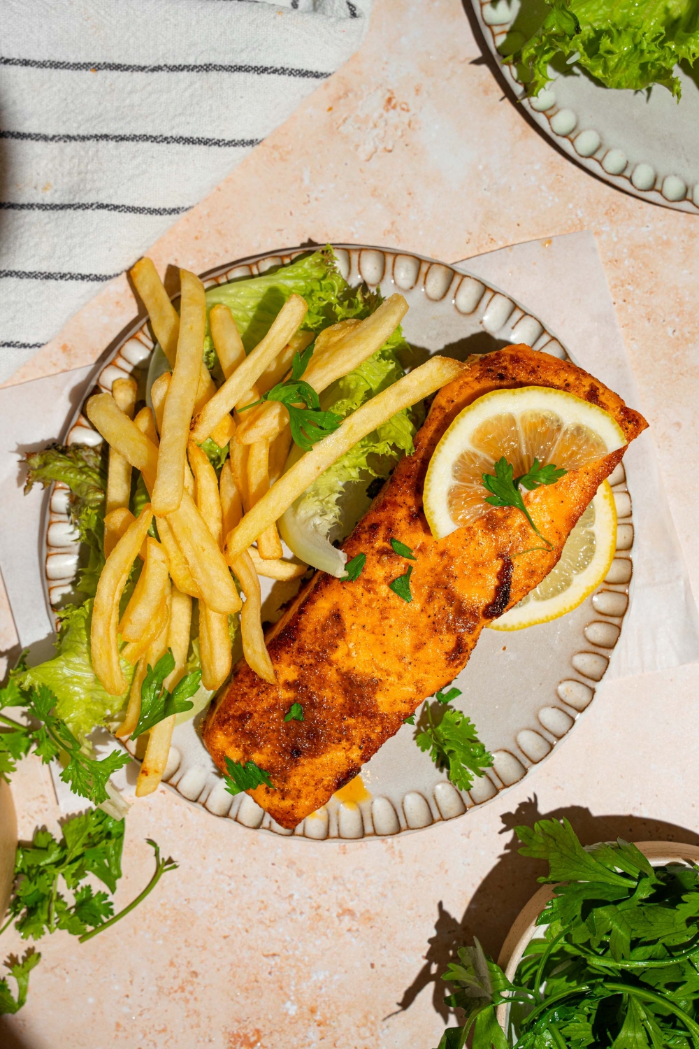 A ceramic plate with frozen air fryer salmon served with lettuce and french fries and garnished with fresh parsley and lemon. The plate is on a tan counter with a white striped napkin.