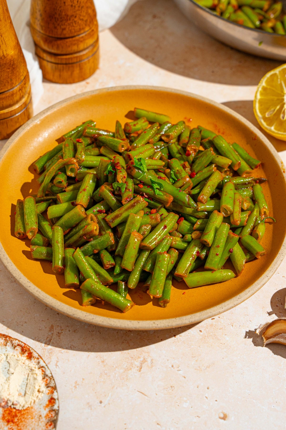 An orange ceramic plate with sautéed frozen green beans tossed in seasonings. The plate is garnished with fresh parsley. The plate is on a tan counter with a salt and pepper mill and small plate of seasonings.