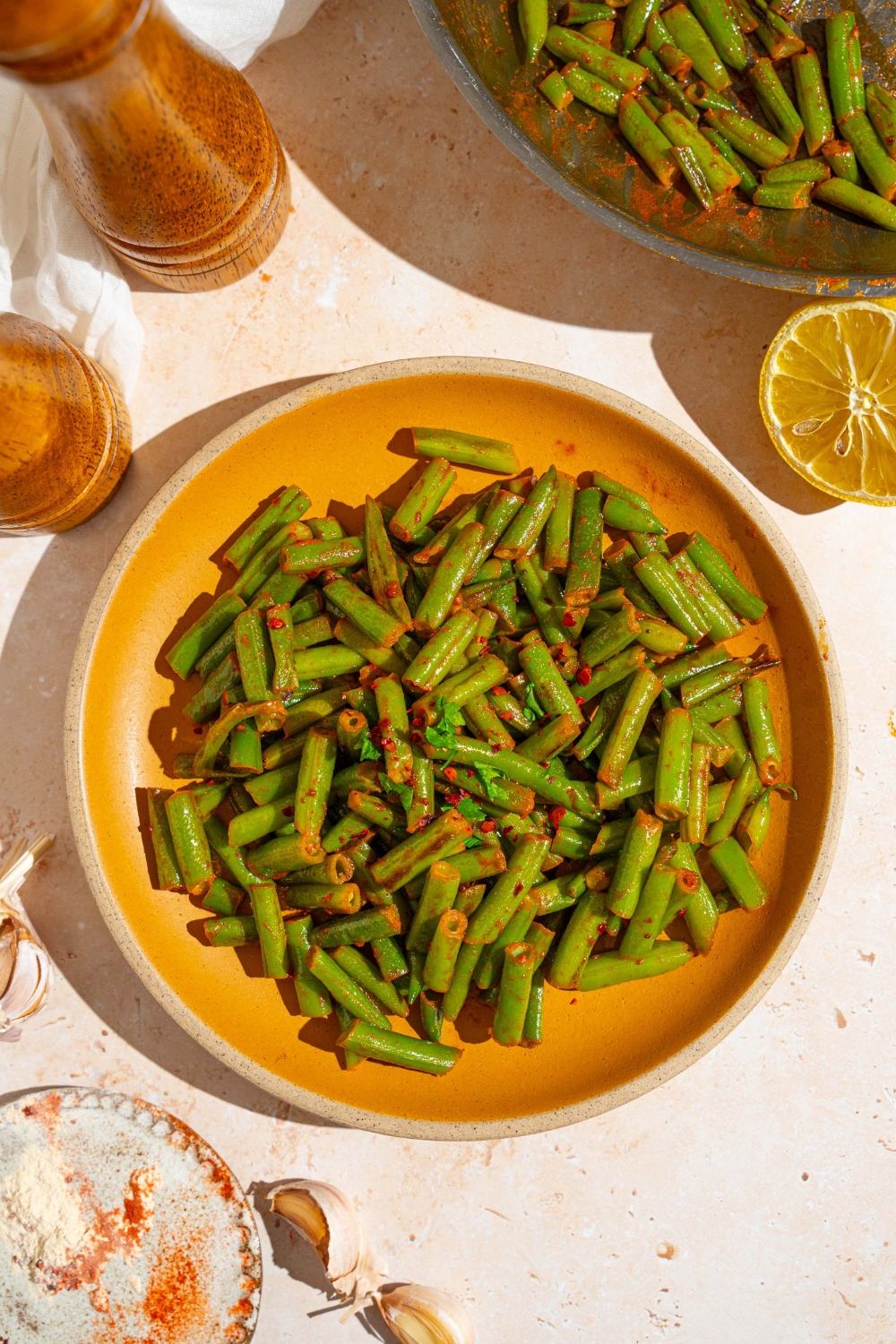 An orange ceramic plate with sautéed frozen green beans tossed in seasonings. The plate is garnished with fresh parsley. The plate is on a tan counter with a salt and pepper mill and small plate of seasonings.