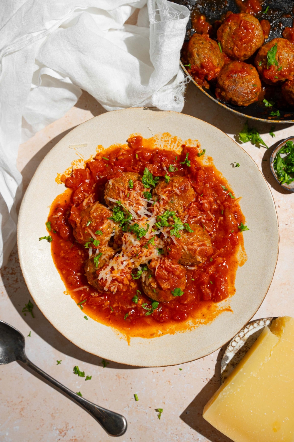 A white plate with frozen meatballs cooked in tomato sauce topped with grated cheese and fresh parsley. The plate is on a tan counter with a spoon and skillet of meatballs.