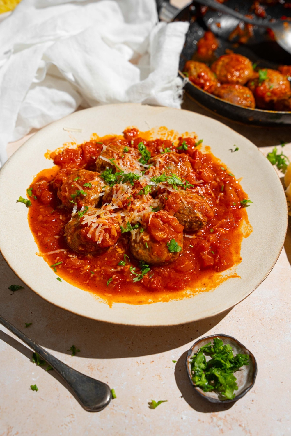 A white plate with frozen meatballs cooked in tomato sauce topped with grated cheese and fresh parsley. The plate is on a tan counter with a spoon and skillet of meatballs.