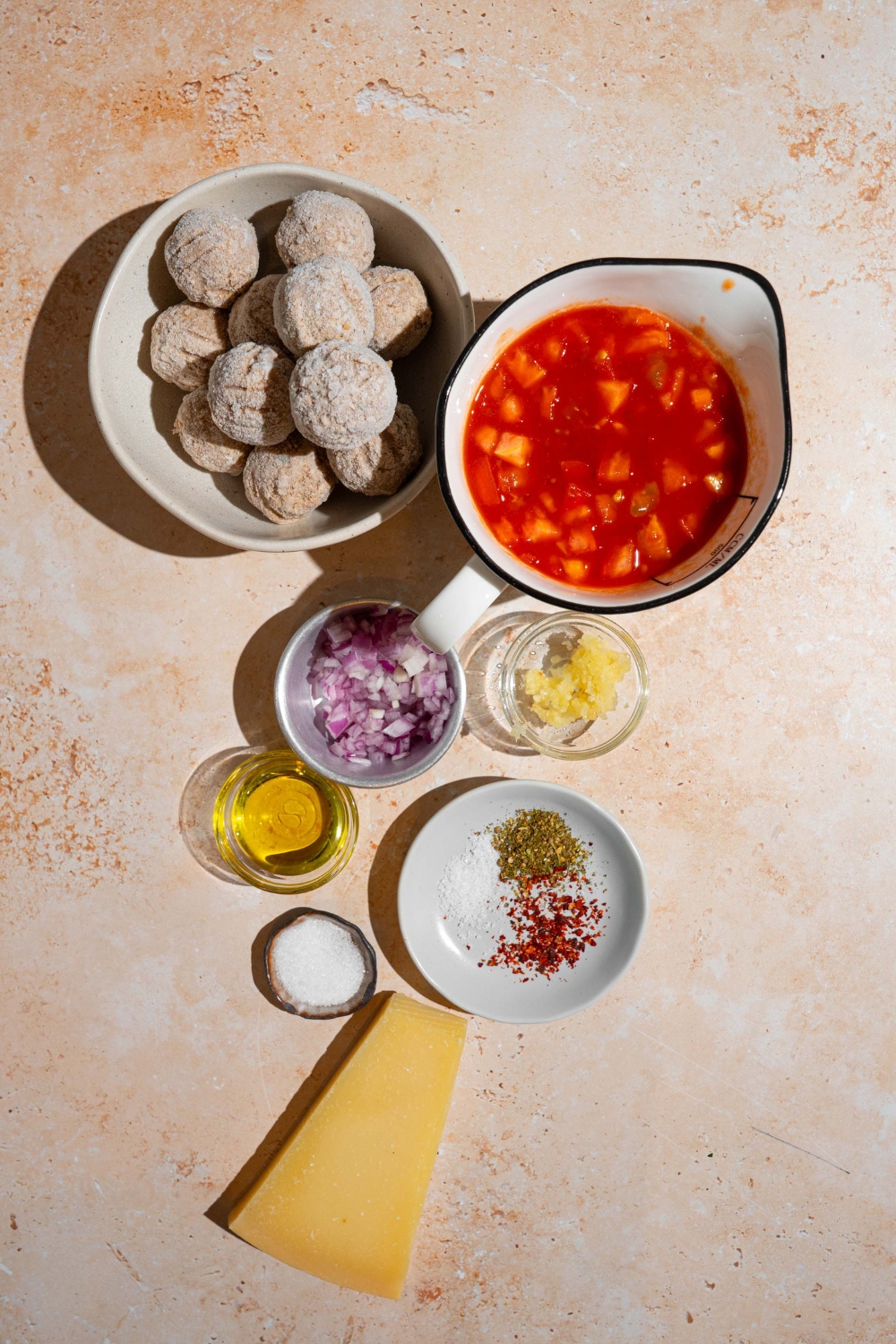 An overhead shot of several bowls in various sizes containing ingredients to make frozen meatballs in tomato sauce including frozen meatballs, diced tomatoes, onion, garlic, parmesan cheese, oil, and seasonings.